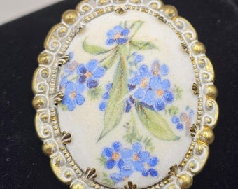 Może przedstawiać: Owalna broszka z pozłacaną metalową ramką i białym porcelanowym środkiem z niebiesko-zielonym wzorem kwiatowym. Środek porcelanowy ma delikatny wzór kwiatowy z kwiatów niezapominajek.