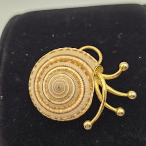 May include: A gold-toned brooch featuring a spiral seashell design with a series of curved, gold-colored accents. The brooch has a natural, cream and brown color palette, and is set against a black background.