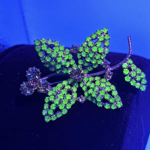 May include: A vintage brooch shaped like a flower, featuring green rhinestone petals and clear rhinestone accents. The brooch has a silver-tone metal stem. The background is a dark blue colour.