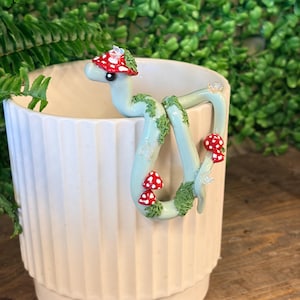 May include: A light green ceramic mug with vertical ridges featuring a sculpted light green snake figurine draped over the rim. The snake is decorated with small red and white toadstool mushrooms, green moss-like textures, and tiny clear crystals.