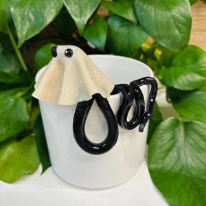 May include: A white ceramic mug featuring a decorative attachment on the rim. The ornament consists of a glossy, cream-colored ghost figure with two small black eyes, connected to a long, smooth, black snake that coils around the mug's exterior.