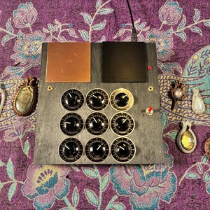May include: A rectangular gray electronic device featuring nine black circular control knobs arranged in a three-by-three grid, a copper square plate, a black square plate, a red toggle switch, and a small red indicator light on the top surface.