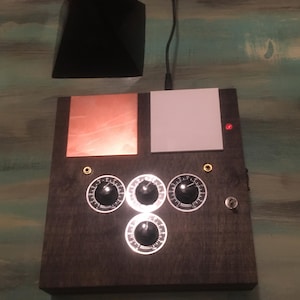 May include: A dark wooden box with four dials, a copper-colored square, and a white square. A black pyramid sits above the box. A red light is on the box. A black cord extends from the box.