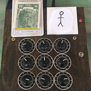 May include: A dark wooden control box featuring nine black circular knobs with white numerical scales from 0 to 100. A 6 of Swords tarot card with a two-dollar bill and a white paper square with a hand-drawn stick figure are attached to the top.