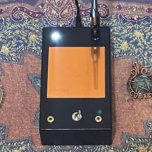 May include: A black electronic device with an orange screen, a silver antenna, and a silver switch. Two wire-wrapped pendants, one with a pink stone and the other with a blue stone, are on either side of the device. The background is a patterned fabric.
