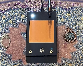 May include: A black electronic device with an orange screen, a silver antenna, and a silver switch. Two wire-wrapped pendants, one with a pink stone and the other with a blue stone, are on either side of the device. The background is a patterned fabric.