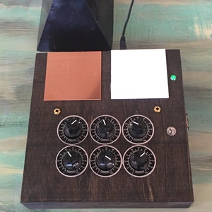 May include: A dark wooden box with six dials, a copper square, and a white square. A black pyramid sits on top. A green light is illuminated on the right side of the box. A black cable extends from the top.