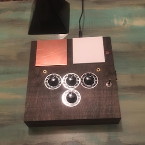 May include: A dark wooden box with copper and white panels, three knobs, and a switch. A black pyramid sits above the box, connected by a wire. The knobs have white markings.