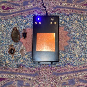 May include: A black rectangular device with a glowing orange square and a blue light, next to four handcrafted gemstone pendants. The pendants feature various stones in copper wire settings, displayed on a patterned fabric.