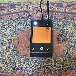 May include: A black electronic device with an orange screen and a small antenna is centered on a patterned textile. Two pendants, one with a pink stone and the other with a green stone, flank the device. The textile has a blue, purple, and gold design.