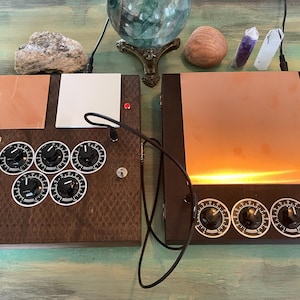 May include: Two dark brown wooden boxes with copper panels and black dials. One box has six dials, the other has three. A black cable connects the boxes. Crystals and a decorative glass sphere are in the background.