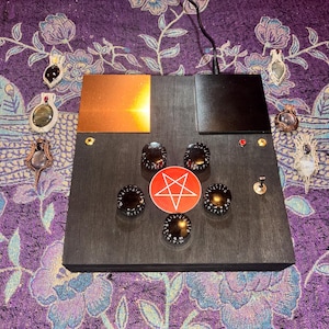 May include: A dark wooden box with a copper plate and black panel. It features six black knobs, a red and white pentagram, and a toggle switch. Several gemstone pendants are arranged around the box on a purple floral patterned fabric.