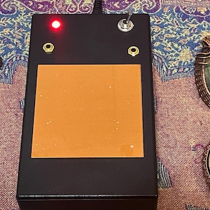 May include: Black rectangular device with an orange square, a red light, and a toggle switch. Two gold-colored eyelets are visible. Decorative wire-wrapped pendants with gemstones are on either side of the device.