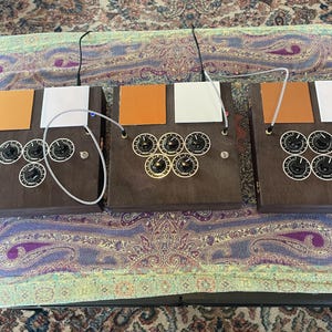 May include: Three dark brown wooden electronic devices with knobs and colored panels. Each device has multiple black knobs with white markings and a silver cable connecting two of them. The panels are orange and white.