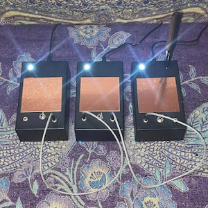May include: Three black electronic devices with copper-colored square panels and bright white lights. Each device has a silver cable and a small switch. One device has a black stylus. The devices are on a purple floral patterned surface.