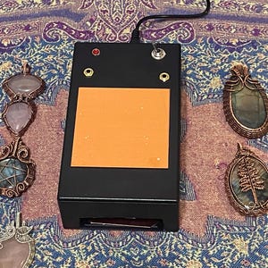 May include: A black device with an orange square, a red light, and a switch, next to several gemstone pendants with copper wire wrapping. The pendants feature various shapes and colors, including pink, blue, and green stones. The items are on a patterned fabric.