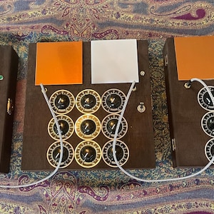 May include: Three brown electronic devices with multiple knobs and switches. Each device has a series of black knobs with white markings and is connected by silver cables. Orange and white squares are on top of each device.