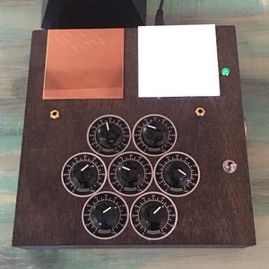 May include: A dark wooden box with a copper square, white square, and seven black knobs with white markings. A green light and a switch are also visible. A black pyramid is on top.