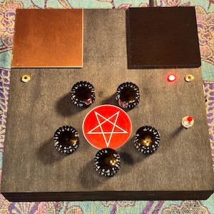 May include: A rectangular dark gray wooden electronic device featuring five black control knobs arranged in a pentagon shape around a red circular sticker with a white pentagram. It includes a copper plate, a black square, and a red LED light.