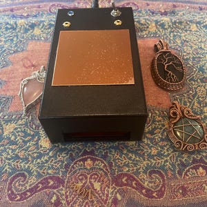 May include: A black electronic device with a copper plate on top, featuring a switch and two small lights. Three handcrafted pendants with gemstones and intricate wirework are also visible, showcasing a variety of shapes and colors. The items are displayed on a patterned fabric.