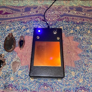 May include: A black electronic device with a copper plate and a blue light, next to four wire-wrapped gemstone pendants. The pendants feature various stones in shades of black, gray, and pink. The background is a patterned textile.