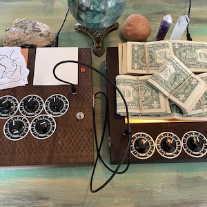 May include: Two dark brown wooden boxes with dials and knobs, connected by a black cable. One box has stacks of dollar bills. The other has a drawing and a white square. Decorative items are in the background.