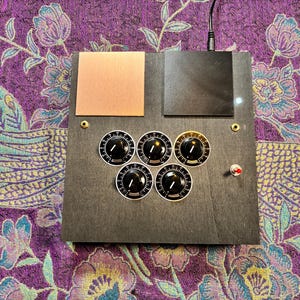 May include: A square black electronic control panel featuring five circular black knobs with white markings arranged in a pentagon pattern, two rectangular plates in copper and black, two small gold-colored ports, and a single red toggle switch.