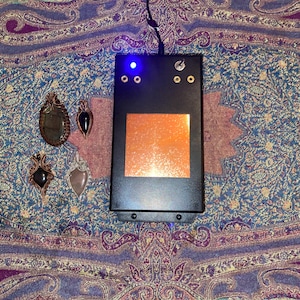 May include: A black rectangular device with a glowing orange square and a blue light, next to four handcrafted gemstone pendants. The pendants feature various stones in copper wire settings, displayed on a patterned fabric.
