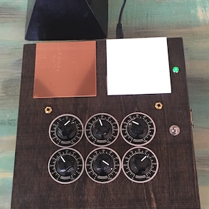 May include: A dark wooden box with six black knobs, a copper square, and a white square. A black pyramid sits above the box. A green light is on the right side of the box.