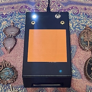 May include: A black device with an orange square, a power switch, and two lights. Four handcrafted pendants with wire-wrapped gemstones in various shapes and colors surround the device. The pendants include rose quartz, labradorite, and other stones.