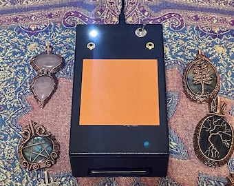 May include: A black device with an orange square, a power switch, and two lights. Four handcrafted pendants with wire-wrapped gemstones in various shapes and colors surround the device. The pendants include rose quartz, labradorite, and other stones.