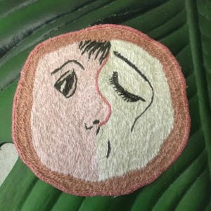 Könnte beinhalten: Ein kreisförmiger gestickter Aufnäher mit einem geteilten Gesichtsmotiv. Die linke Seite ist hellrosa mit einem dunklen Auge und einer Augenbraue, während die rechte Seite cremeweiß mit einem geschlossenen Auge und langen Wimpern ist. Der Rand ist braun.