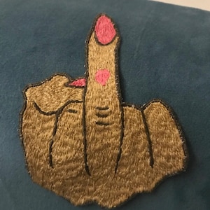 Può includere: Toppa ricamata raffigurante una mano che fa il dito medio. La toppa è beige con contorni neri e smalto rosa. Lo sfondo è blu-azzurro.
