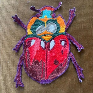 Può includere: Toppa ricamata a forma di scarabeo colorato. La toppa presenta un corpo rosso con dettagli bianchi, testa arancione e verde, e zampe e antenne viola. Progettata per l'applicazione su abbigliamento o accessori.