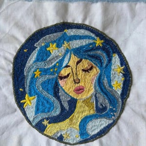 Può includere: Toppa ricamata con un design celeste. Raffigura una donna con capelli blu e stelle dorate, su uno sfondo circolare blu scuro. Il viso della donna è giallo con gli occhi chiusi e le labbra rosa. La toppa è su un tessuto bianco.