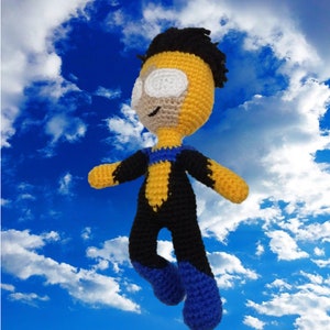 May include: A crocheted plush toy of a superhero wearing a yellow and black costume with blue boots. The toy is posed with arms outstretched and legs bent, as if flying.