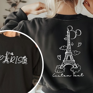 May include: Black sweatshirt with a white graphic of the Eiffel Tower, Mickey Mouse ears, and hearts. The text "Paris" and Mickey Mouse figures are on the front of the sweatshirt. The text "Custom Text" is on the back of the sweatshirt.