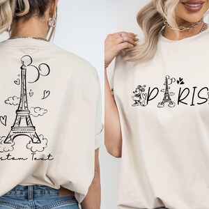 May include: Two white t-shirts with a black graphic design. One shirt features a cartoon Eiffel Tower with hearts and clouds. The other shirt features the word "PARIS" with a cartoon Eiffel Tower and Mickey Mouse characters.