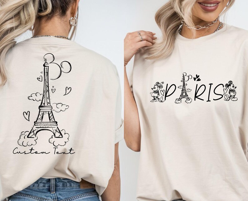 May include: Two white t-shirts with a black graphic design. One shirt features a cartoon Eiffel Tower with hearts and clouds. The other shirt features the word "PARIS" with a cartoon Eiffel Tower and Mickey Mouse characters.