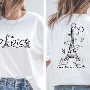 May include: White t-shirt with a black and white graphic of Mickey Mouse characters and the Eiffel Tower with the word "PARIS" printed on the front. The back of the shirt has a black and white graphic of the Eiffel Tower with clouds and hearts and the text "Custom Text".