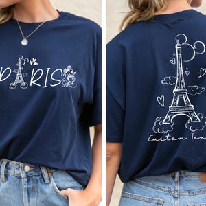 May include: A navy blue t-shirt with a white graphic of Mickey Mouse and Minnie Mouse in front of the Eiffel Tower with the word "PARIS" printed below. The back of the shirt has a white graphic of the Eiffel Tower with clouds and hearts and the text "Custom Text".