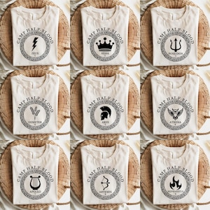 May include: Nine white t-shirts with black circular designs featuring the names of Greek gods and goddesses. Each design includes the text "Camp Half-Blood" and the god or goddess's name. The gods and goddesses featured are Zeus, Hera, Poseidon, Demeter, Ares, Athena, Apollo, Artemis, and Hephaestus.