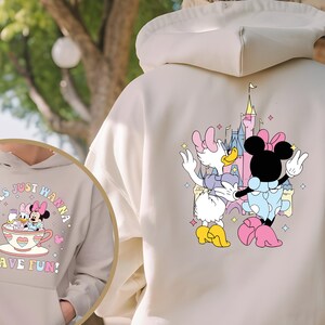 May include: A light beige hoodie with a graphic print of two Disney characters, Minnie Mouse and Daisy Duck, in front of Cinderella's Castle. The text "Girls Just Wanna Have Fun!" is printed on the front of the hoodie.