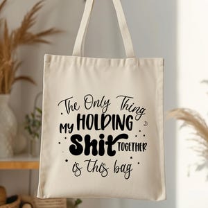 May include: A white canvas tote bag with black text that reads "The Only Thing My Holding Shit Together Is This Bag."