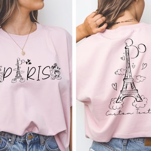 May include: Pink t-shirt with a black and white graphic of Mickey Mouse and Minnie Mouse in front of the Eiffel Tower with the word "PARIS" printed below. The back of the shirt has a black and white graphic of the Eiffel Tower with clouds and hearts. The text "Custom Text" is printed below the Eiffel Tower.
