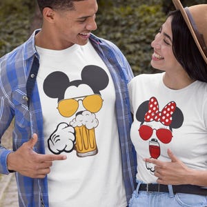May include: Two people wearing white t-shirts with cartoon character designs. One shirt features Mickey Mouse with sunglasses holding a beer, the other features Minnie Mouse with sunglasses and a drink. The couple is smiling.
