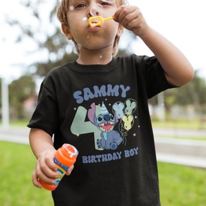 K&ouml;nnte beinhalten: Ein schwarzes T-Shirt mit einer blauen Stitch-Figur, die Ballons h&auml;lt. Das Shirt tr&auml;gt den Schriftzug "Sammy 4 Birthday Boy" in einer bunten Schrift.