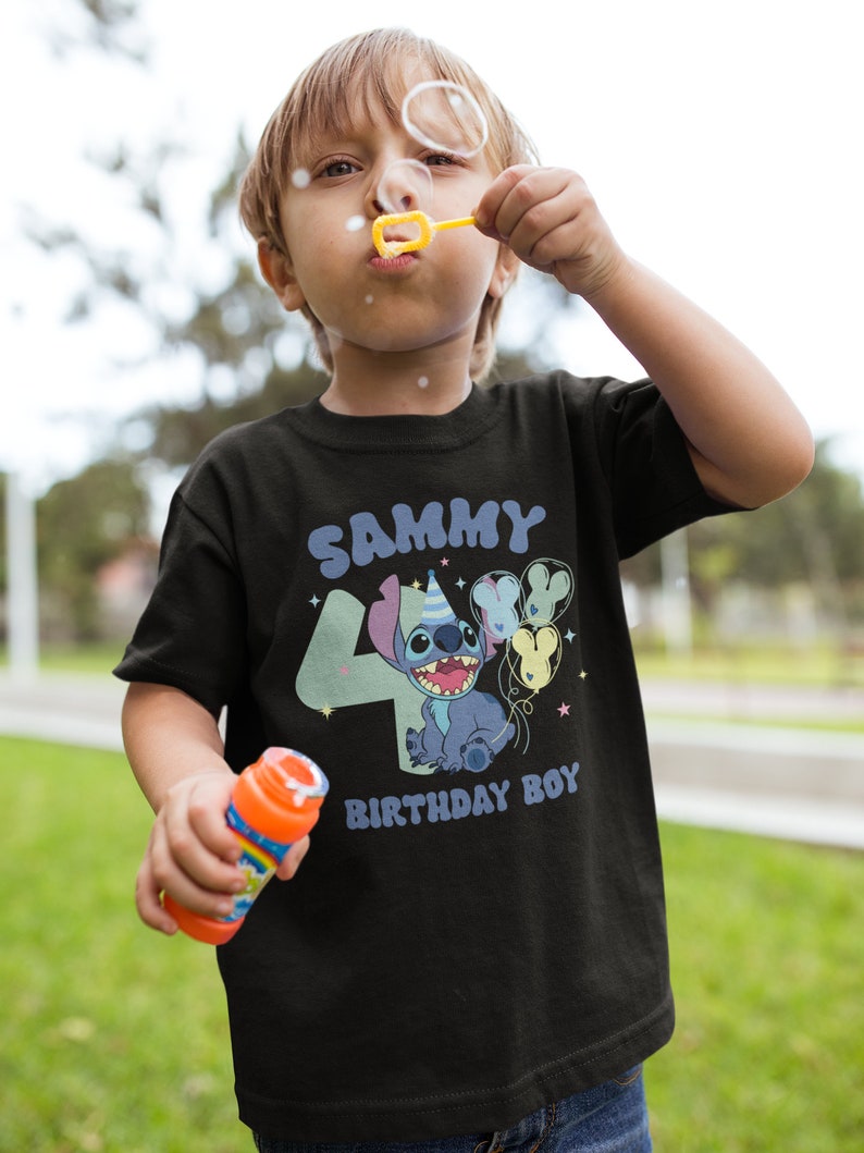 K&ouml;nnte beinhalten: Ein schwarzes T-Shirt mit einer blauen Stitch-Figur, die Ballons h&auml;lt. Das Shirt tr&auml;gt den Schriftzug "Sammy 4 Birthday Boy" in einer bunten Schrift.