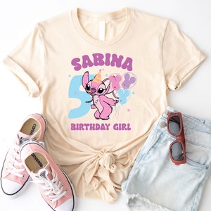K&ouml;nnte beinhalten: Ein hellbeiges T-Shirt mit einer lilafarbenen Grafik eines Comic-Charakters, der Ballons h&auml;lt. Der Text "SABINA BIRTHDAY GIRL" ist auf das T-Shirt gedruckt.