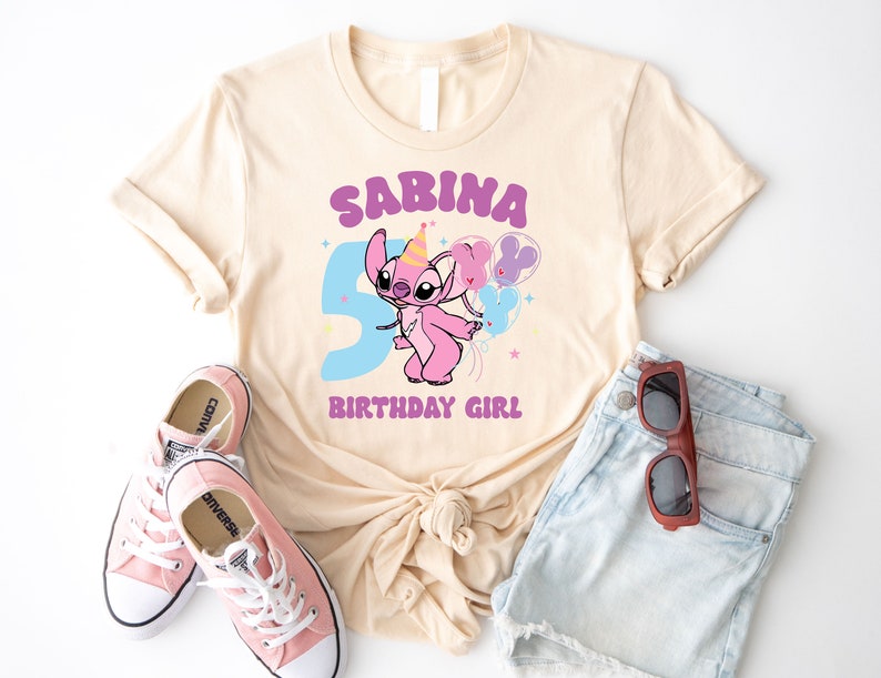 K&ouml;nnte beinhalten: Ein hellbeiges T-Shirt mit einer lilafarbenen Grafik eines Comic-Charakters, der Ballons h&auml;lt. Der Text "SABINA BIRTHDAY GIRL" ist auf das T-Shirt gedruckt.